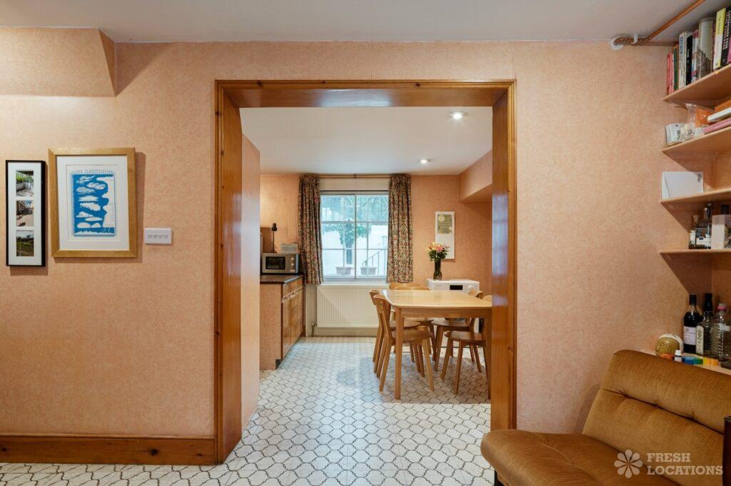 Retro shoot locations: step inside timeless homes for standout shoots 11 Georgian-style kitchen of gingernut, a london townhouse shoot location featuring 1960s interiors. The space features peach-toned textured wallpaper, retro wood trim, and patterned hexagonal floor tiles. A light wood dining table with matching chairs sits near a window with floral curtains.