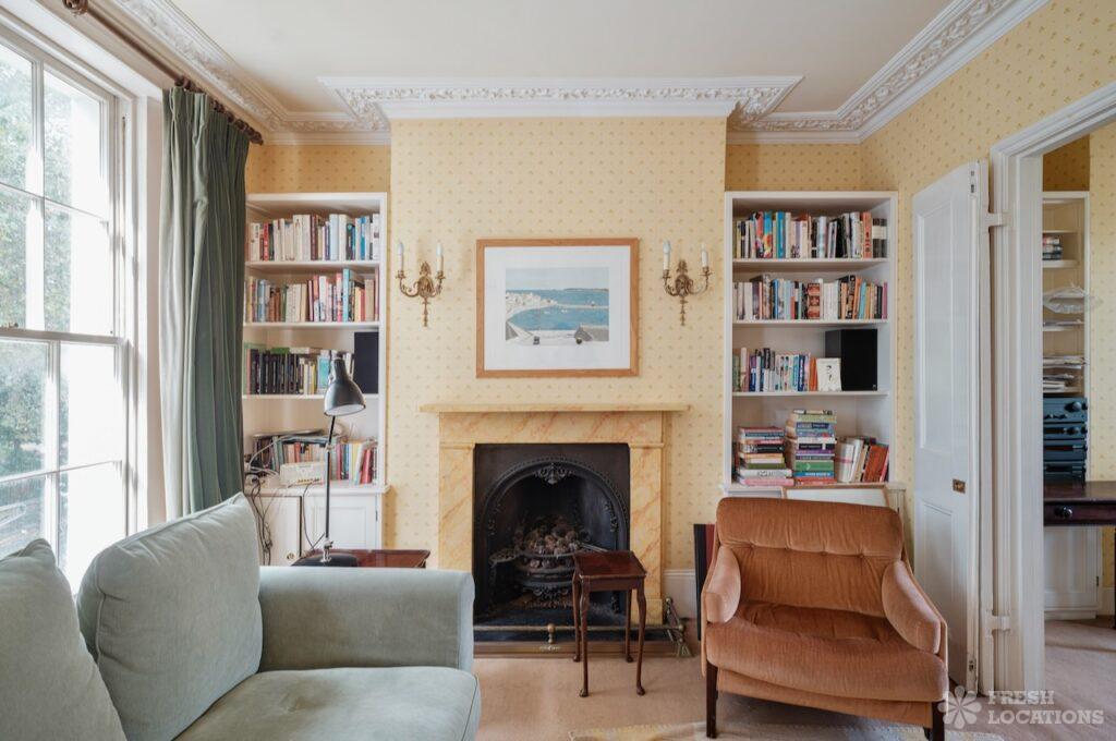 Retro shoot locations: step inside timeless homes for standout shoots 10 Georgian-style living room of gingernut, a london townhouse shoot location featuring 1960s interiors. The space includes a yellow patterned wallpaper, a vintage marble fireplace with a cast iron insert, flanked by built-in bookshelves filled with books. A seaside art print and antique-style brass wall sconces add to the mid-century aesthetic. Furnishings include a green velvet sofa and a retro rust armchair, with decorative ceiling cornicing and natural light from a large sash window.