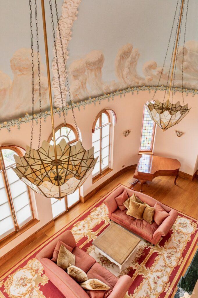Retro shoot locations: step inside timeless homes for standout shoots 9 Lavish rococo-style living room in a north west london shoot location called rococo château. Viewed from above, the room features vaulted ceilings painted with clouds and adorned with intricate plaster reliefs and floral detailing, grand arched windows, and dramatic pendant chandeliers. Below, two blush pink velvet sofas sit on an ornate red and gold rug beside a polished wood baby grand piano and light wood floors, creating an opulent 18th-century french-inspired aesthetic.
