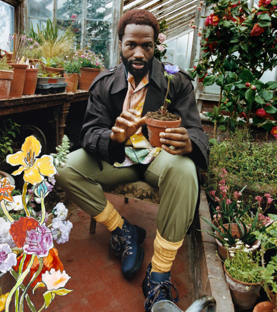 English actor sope dirisu seated in a rustic greenhouse at 'bedmond', a countryside shoot location in hertfordshire, for the highgrove x burberry campaign. He wears layered burberry pieces; an oversized trench, printed shirt, olive trousers, and yellow socks with blue hiking boots, while holding a potted flower. Surrounded by blooming plants and painted floral illustrations, the setting blends heritage fashion with an organic, pastoral aesthetic.