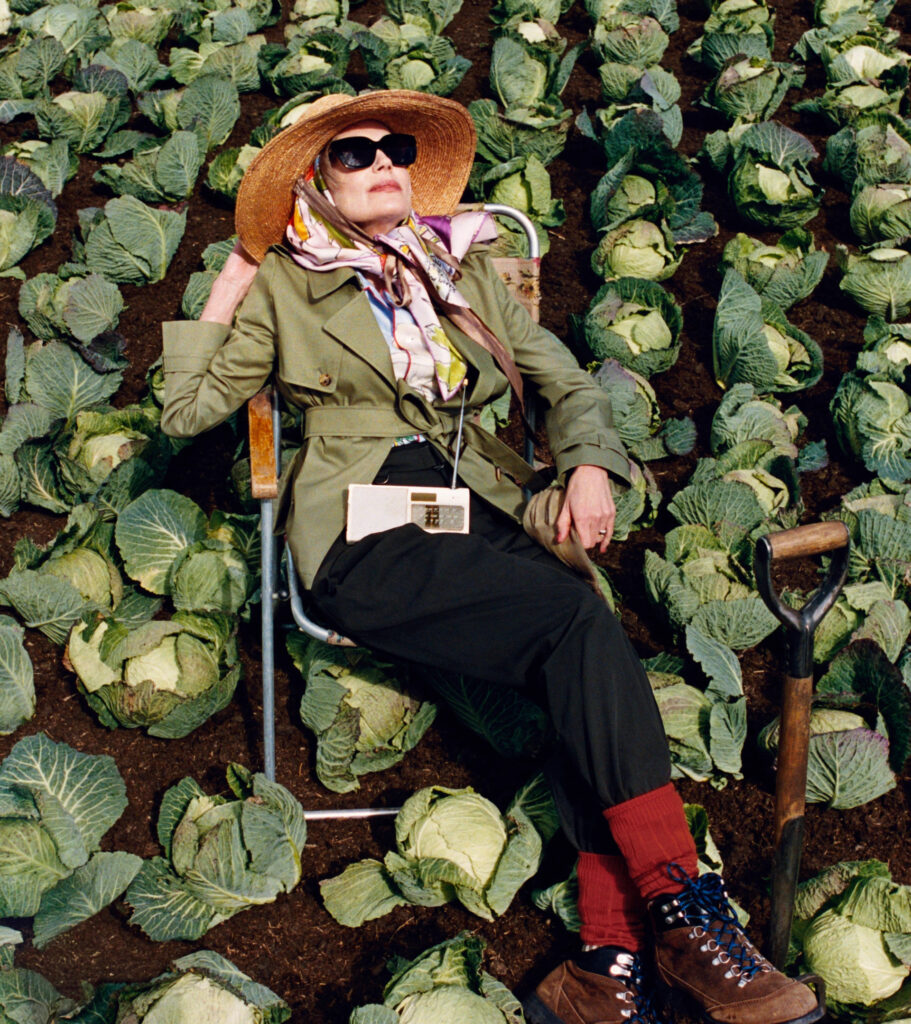 American actress elizabeth mcgovern reclining in a fold-out chair among rows of cabbages in a cultivated field at 'bedmond', a countryside shoot location in hertfordshire, for the highgrove x burberry campaign. She wears a green trench coat, floral silk scarf, oversized sunglasses, and a straw sun hat, styled with hiking boots and red socks. A white burberry clutch is fastened around her waist, and a garden spade stands nearby, blending classic british fashion with whimsical rural life.