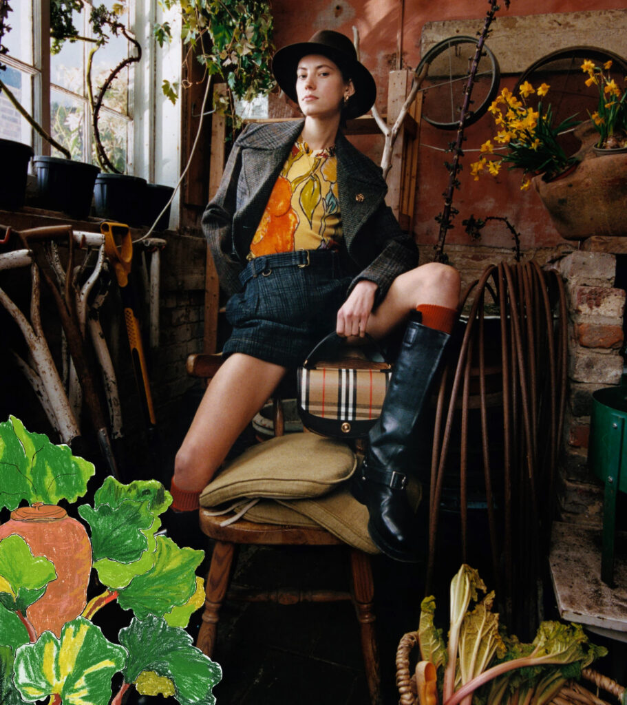 A model sits confidently in a rustic potting shed at bedmond, a countryside shoot location in hertfordshire, for the highgrove x burberry campaign. She wears a wide-brim hat, a vintage-inspired tweed jacket, a bold floral shirt, and tailored shorts, styled with knee-high black boots and rust-coloured socks. A burberry check handbag rests on a chair beneath her, surrounded by gardening tools, daffodils, and worn brick walls—evoking a modern countryside elegance.