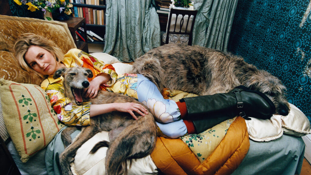 English actress laura carmichael lounges on a bed at bedmond wearing bold floral burberry silk pyjamas and tall leather boots, flanked by two large irish wolfhounds. One dog is sprawled across her legs, the other nestled in her arms mid-yawn. The room features traditional british decor—patterned cushions, embroidered linens, and a wallpapered wall with blue floral motifs—adding to the intimate, cozy mood of the shoot.
