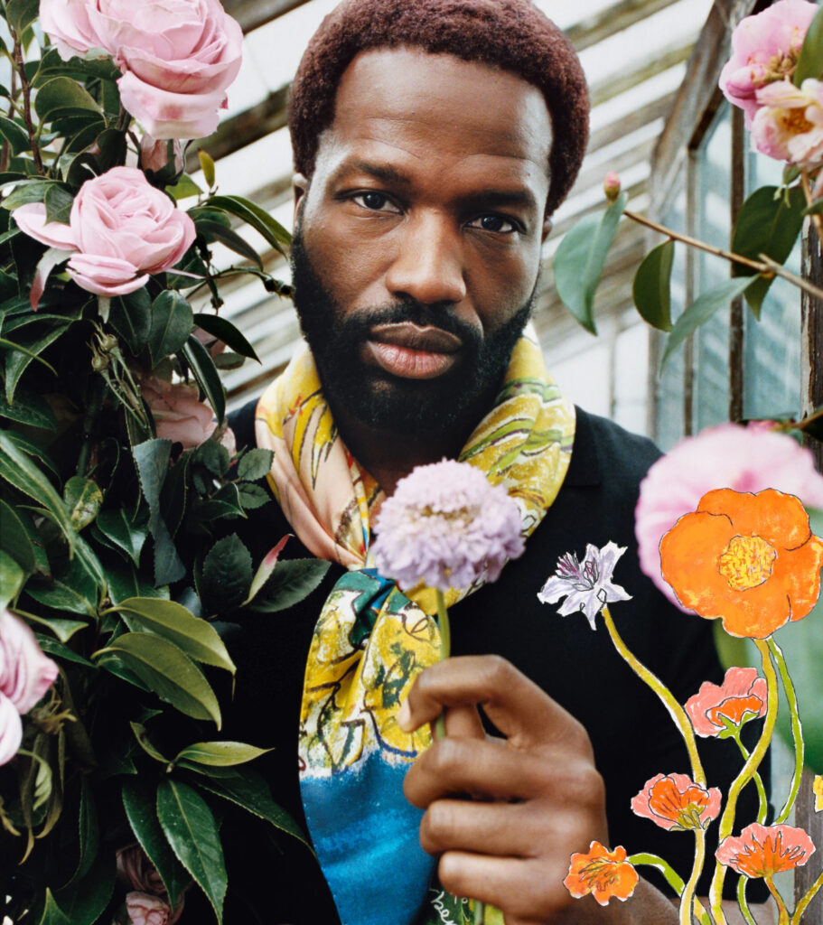 Close up portrait english actor sope dirisu holding a flower in a rustic greenhouse at 'bedmond', a countryside shoot location in hertfordshire, for the highgrove x burberry campaign. He wears a burberry neck scarf with an abstract floral print and black t-shirt. Surrounded by blooming pink roses and painted floral illustrations, the setting blends heritage fashion with an organic, pastoral aesthetic.