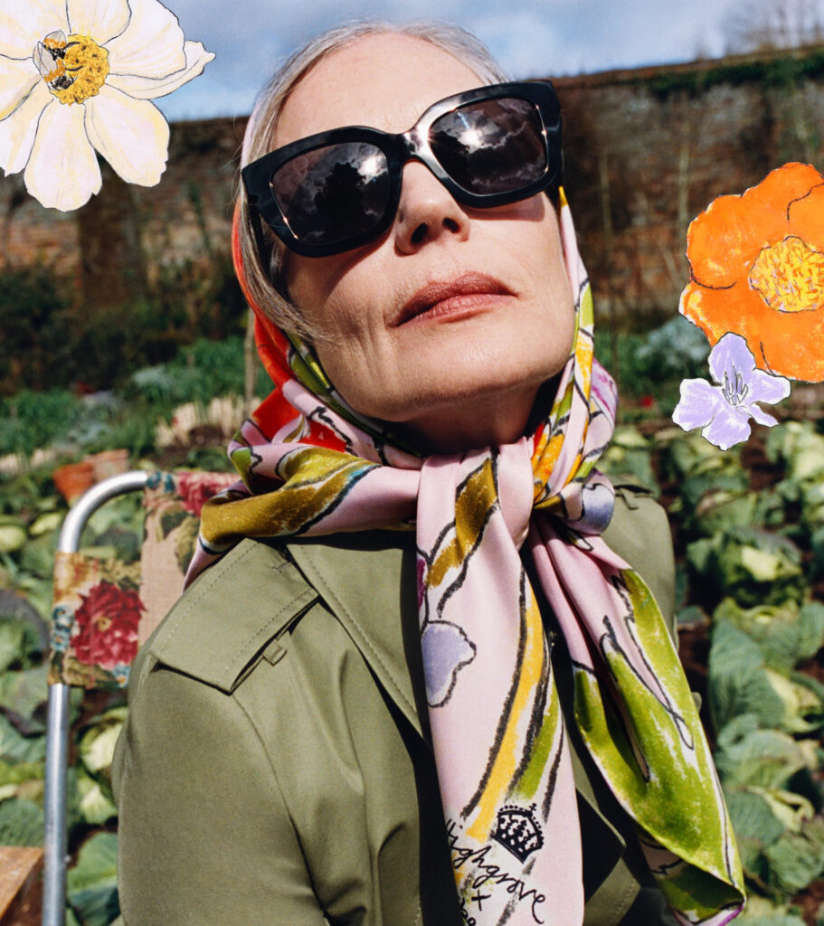 A close up portrait of american actress elizabeth mcgovern seated in a fold-out chair among rows of cabbages in a cultivated field at 'bedmond', a countryside shoot location in hertfordshire, for the highgrove x burberry campaign. She wears a green trench coat, floral silk head scarf and oversized sunglasses. You can see a brick wall in the back ground, blending classic british fashion with whimsical rural life.