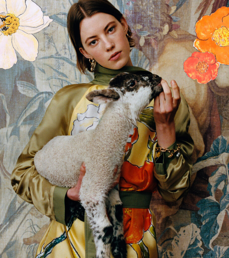A female model holds a black speckled lamb in front of a fabric backdrop that had a faded abstract print of plants, fish and a man. The model wears a long-sleeved silk dress with an abstract print. Photographed at 'bedmond', a countryside shoot location in hertfordshire, for the highgrove x burberry campaign.